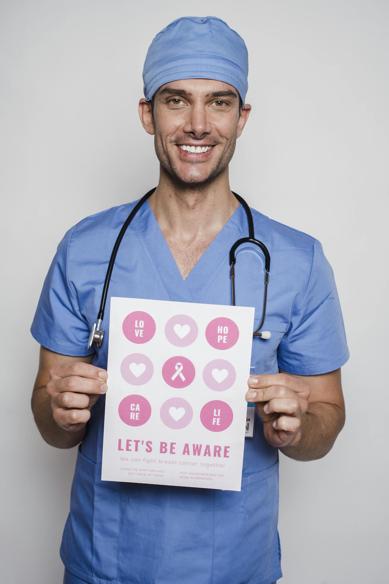 Smiling doctor in blue scrubs holding a breast cancer awareness poster with pink symbols.